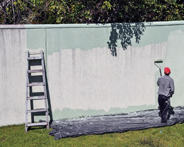 Imagem: Fotografia. Um homem está de costas e segurando um rolo de tinta e pintando uma parede de verde-claro. Abaixo dele há uma lona preta estendida no chão.   Fim da imagem.