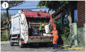 Imagem: Fotografia 1. Uma pessoa com uniforme laranja e botas azuis está jogando um saco de lixo na parte traseira de um caminhão de lixo. Ao fundo, casas e árvores.  Fim da imagem.