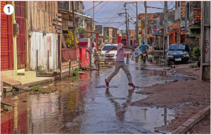 Imagem: Fotografia 1. No centro, uma pessoa está andando sobre uma rua com água. Atrás há carros e em volta, construções e postes.   Fim da imagem.