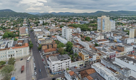 Imagem: Fotografia. Vista aérea de uma cidade com ruas asfaltadas, veículos, prédios, árvores e casas. Fim da imagem.