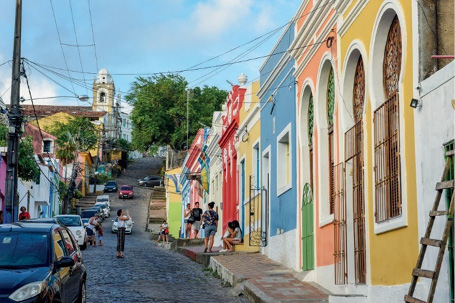 Imagem: Fotografia. No centro há carros em uma rua estreita. À direita, construções coloridas, uma ao lado da outra.  Fim da imagem.