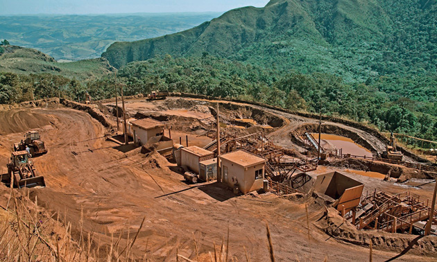 Imagem: Fotografia. Vista aérea de tratores sobre terra escura. Ao lado há duas construções pequenas e atrás, um morro com escavações. Ao fundo, morros e árvores. Fim da imagem.