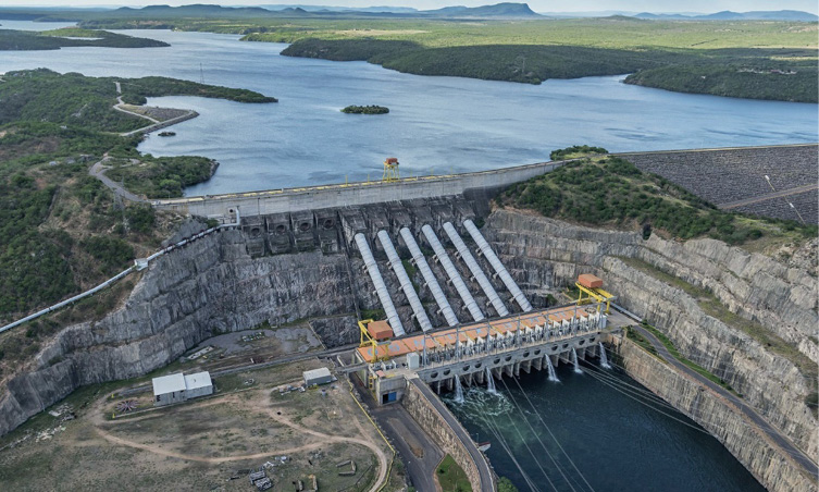 Imagem: Fotografia. Vista aérea de um lago. Ao lado há uma barragem com canos grossos e grandes. Ao fundo, árvores. Fim da imagem.