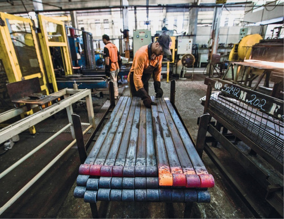 Imagem: Fotografia. Um homem com boné, avental e luvas pretas e uniforme laranja está com as mãos sobre filetes de metal empilhados. Ao fundo, máquinas e pessoas. Fim da imagem.