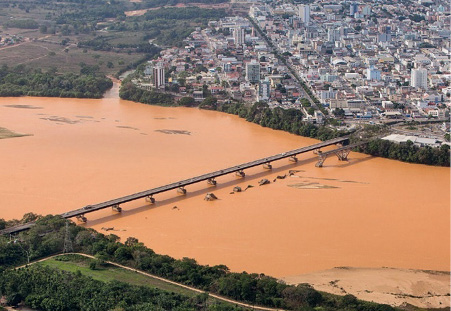 Imagem: Fotografia. No centro há um rio com água marrom. Sobre ele há uma ponte e na margem, vários prédios, casas e ruas. Fim da imagem.