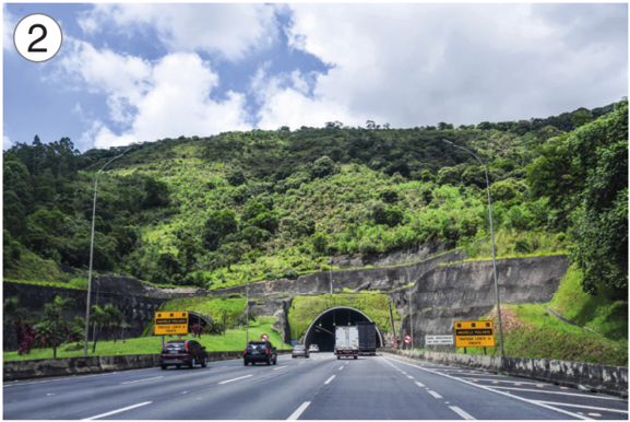 Imagem: Fotografia 2. Carros sobre uma estrada e no centro há um túnel em um morro com árvores. Fim da imagem.