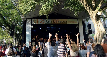 Imagem: Fotografia. Várias pessoas estão de costas e algumas estão com as mãos levantadas. Na frente delas há uma faixa com a informação: 22ª FESTA DO IMIGRANTE. Abaixo há pessoas sobre um palco com as mãos para cima. Em volta há árvores. Fim da imagem.