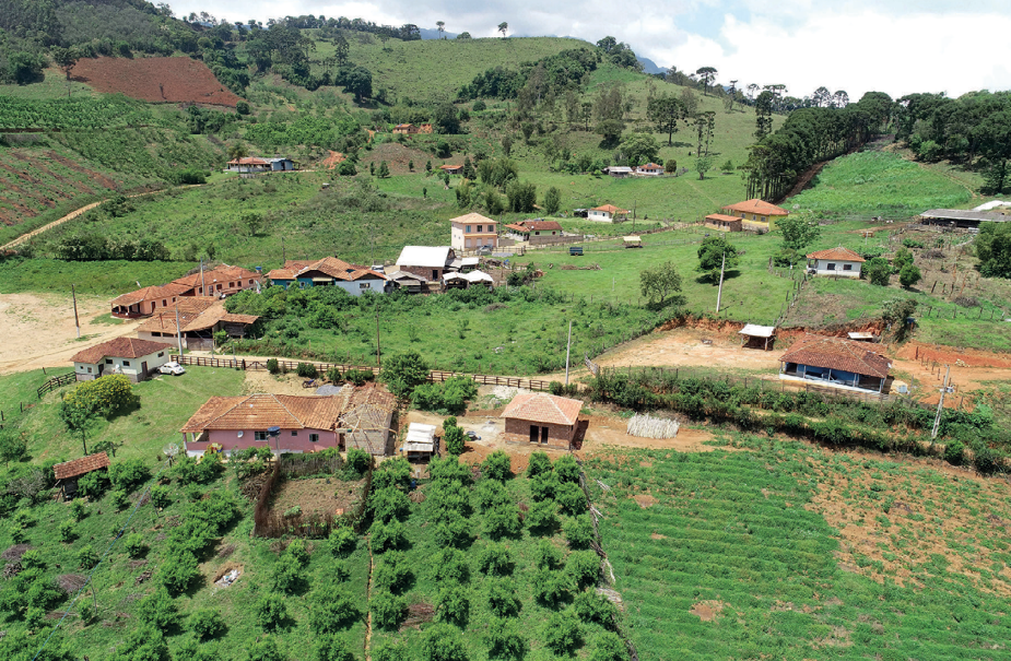 Imagem: Fotografia. Vista aérea de casas espalhadas em um campo. Ao lado de uma casa há uma plantação e ao fundo, morros e árvores. Fim da imagem.