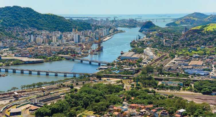 Imagem: Fotografia. No centro há um rio largo e extenso. Sobre ele há três pontes e nas margens há árvores e construções. Ao fundo, morros com árvores em volta. Fim da imagem.