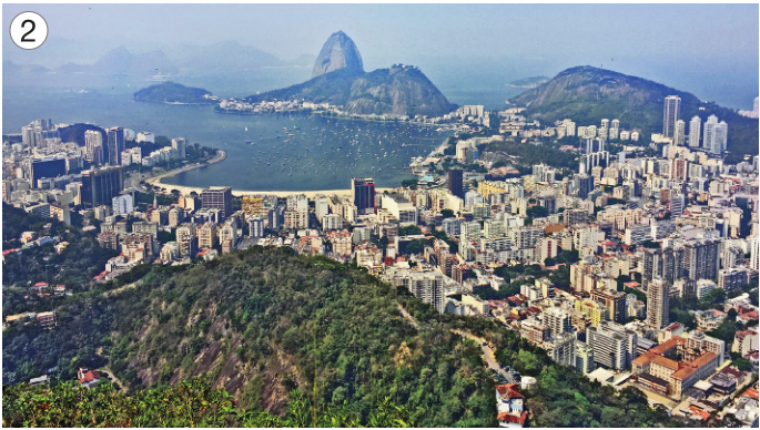 Imagem: Fotografia 2. Vista aérea de um morro cercado de construções. Ao fundo, outro morro cercado pelo mar.  Fim da imagem.