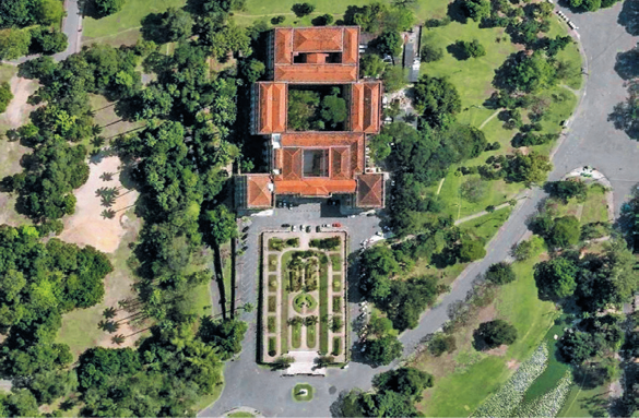 Imagem: Fotografia. Vista de cima de uma construção grande com telhado laranja em volta de árvores. Na frente há uma praça extensa; à esquerda, várias árvores e à direita, um pavimento ao lado de um lago com pedrisco em volta.  Fim da imagem.