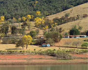 Imagem: Fotografia. No centro há um lago e na margem, poucas casas espalhadas entre árvores. Ao fundo, morros e mais árvores.  Fim da imagem.