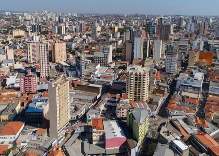 Imagem: Fotografia. Vista aérea de várias construções e prédios entre ruas. Fim da imagem.