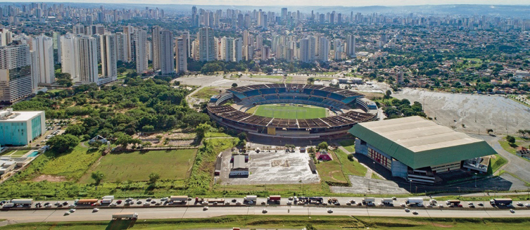 Imagem: Fotografia. Vista aérea de um campo de futebol com uma construção circular em volta. Ao lado há várias árvores e uma estrada com automóveis. Ao fundo, prédios e casas.  Fim da imagem.