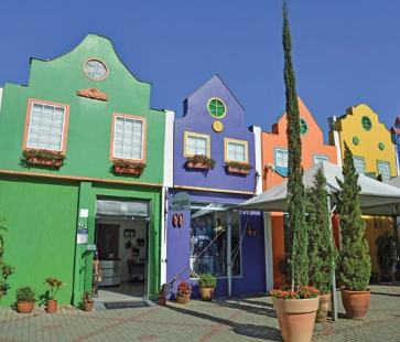 Imagem: Fotografia. Construções com paredes coloridas, lado a lado. Na frente há vasos com plantas.  Fim da imagem.