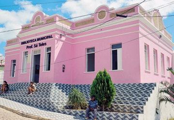 Imagem: Fotografia. Três pessoas estão sentadas em uma escadaria, na frente de uma construção grande com parede rosa. Acima, uma placa com a informação: BIBILIOTECA MUNICIPAL.  Fim da imagem.