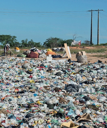 Imagem: Fotografia. Muito lixo empilhado. Ao fundo, árvores. Fim da imagem.