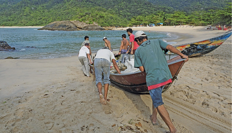 Imagem: Fotografia. Sete homens estão empurrando um barco em direção a um rio. Ao fundo, árvores. Fim da imagem.