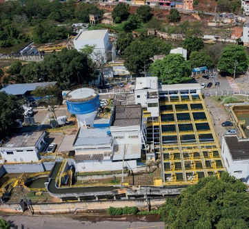 Imagem: Fotografia. Vista aérea de uma estação de tratamento de água. À esquerda, construção grande e à direita, vários tanques com água. Em volta há árvores e ruas. Fim da imagem.