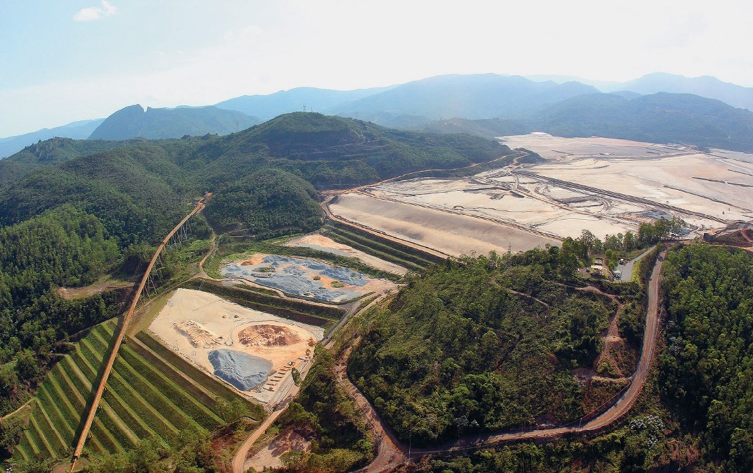 Imagem: Fotografia. Vista aérea de um terreno plano com terra clara no topo de um morro. Em volta há vários morros cobertos de árvores. Fim da imagem.