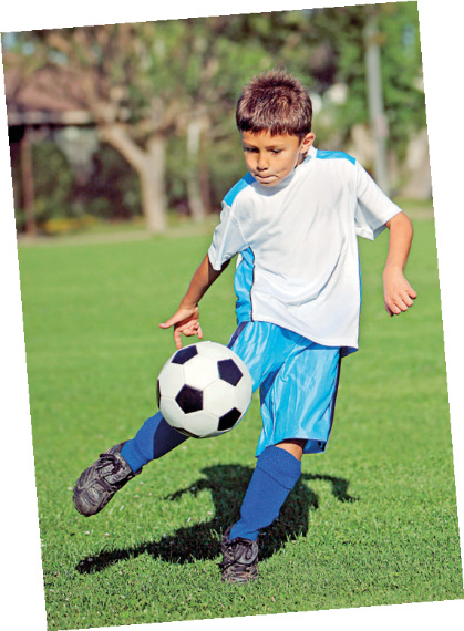 Imagem: Fotografia. Um menino com cabelo castanho e curto, camiseta branca, bermuda azul e chuteiras pretas está chutando uma bola de futebol. Fim da imagem.