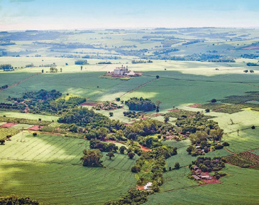 Imagem: Fotografia. Vista aérea de plantações. No centro há casas espalhadas e árvores. Fim da imagem.