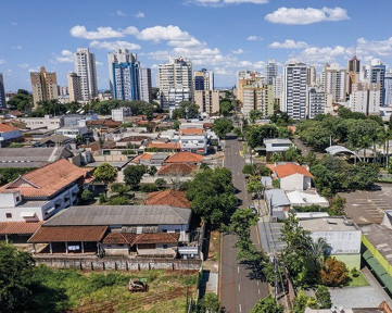 Imagem: Fotografia. Vista aérea de uma rua com árvores em volta. Nas laterais há casas e ao fundo, prédios. Fim da imagem.