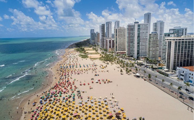 Imagem: Fotografia. Vista aérea de várias pessoas na orla da praia. À direita, vários prédios. Fim da imagem.