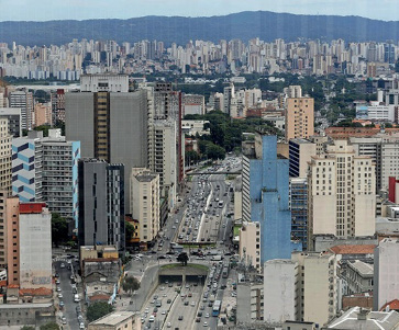 Imagem: Fotografia. Vista aérea de uma avenida com automóveis. Nas laterais há vários prédios.  Fim da imagem.