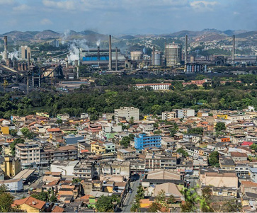 Imagem: Fotografia. Vista aérea de uma cidade com várias construções. No centro há árvores e ao fundo, usinas e fumaça. Fim da imagem.