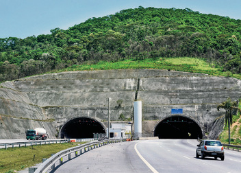 Imagem: Fotografia. Duas estradas asfaltadas. Ao fundo, dois túneis embaixo de morros verdes. Fim da imagem.