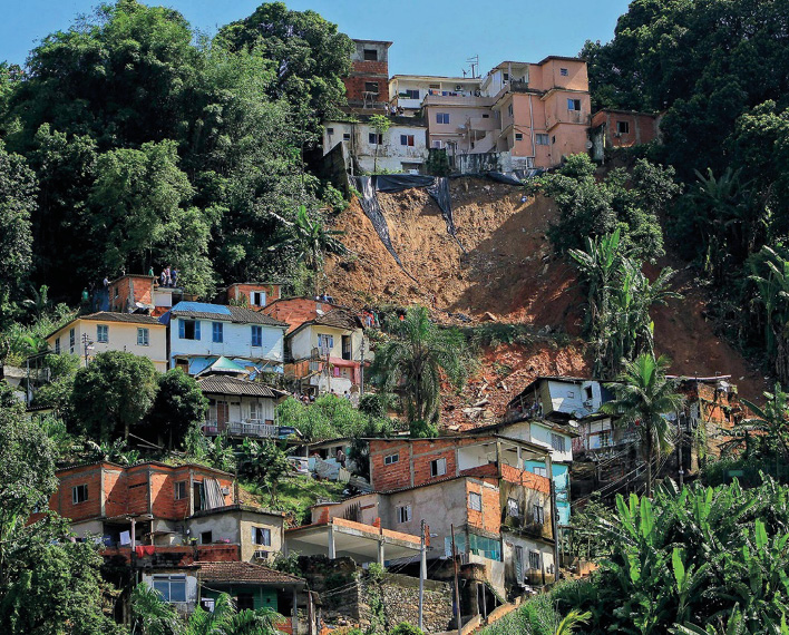 Imagem: Fotografia. Construções sobre a encosta de um morro. No centro há um deslizamento e em volta, várias árvores. Fim da imagem.