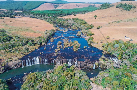 Imagem: Fotografia. Vista aérea de uma queda d’água. Em volta há plantas e árvores e ao fundo, morros. Fim da imagem.