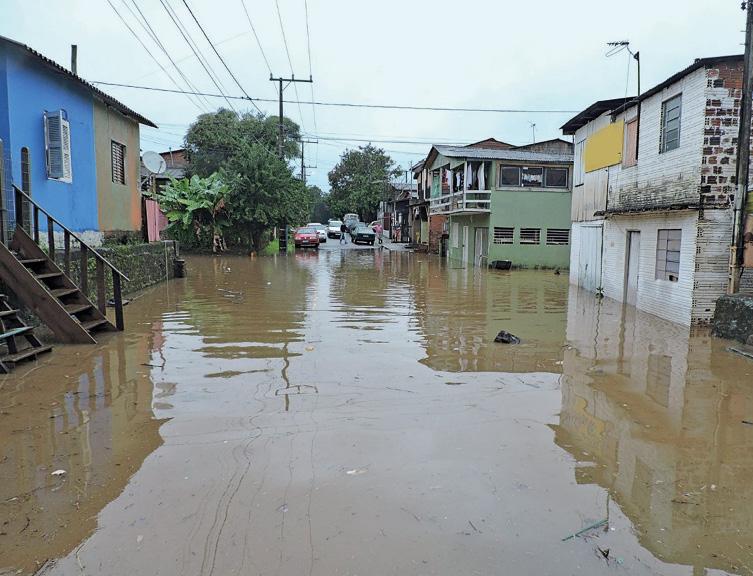 Imagem: Fotografia. Uma rua inundada com água escura. Nas laterais há casas e árvores. Fim da imagem.