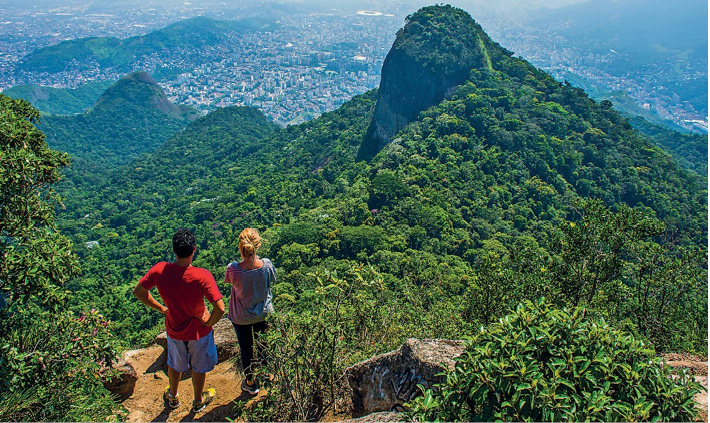 Imagem: Fotografia. Um casal está parado no topo de um morro. Abaixo deles há um morro coberto de árvores e ao fundo, uma cidade. Fim da imagem.