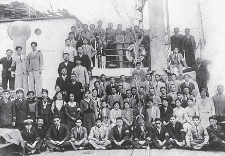 Imagem: Fotografia em preto e branco. Várias pessoas orientais estão aglomeradas em um navio e olhando para frente. Fim da imagem.