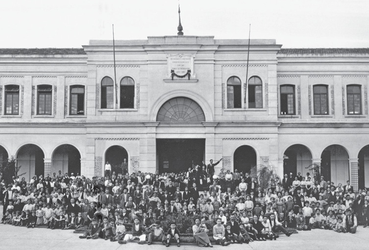 Imagem: Fotografia em preto e branco. Uma multidão está sentada na frente de uma construção grande com dois andares.  Fim da imagem.