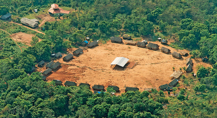 Imagem: Fotografia. Vista aérea de ocas em volta de um galpão grande com telhado. Ao redor há várias árvores. Fim da imagem.