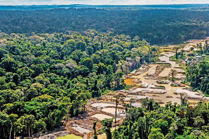 Imagem: Fotografia. Vista aérea de uma floresta. No centro há uma área desmatada com várias poças d’água.  Fim da imagem.