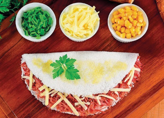 Imagem: Fotografia. Uma tapioca branca com recheio de carne e queijo. Ao lado há recipientes com cebolinha, queijo e milho. Fim da imagem.