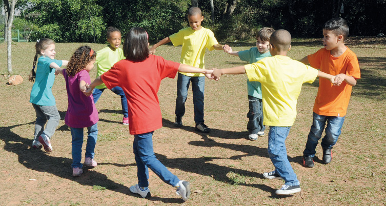 Imagem: Fotografia. Crianças com roupas coloridas estão de mãos dadas e formando uma roda. Ao fundo, árvores. Fim da imagem.