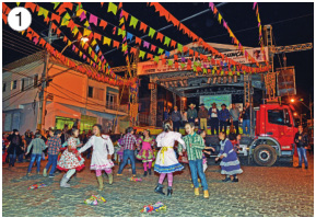 Imagem: Fotografia 1. Crianças com roupas coloridas estão sorrindo e dançando. Atrás delas há pessoas sobre um palco e em volta há várias bandeirinhas coloridas penduradas.  Fim da imagem.