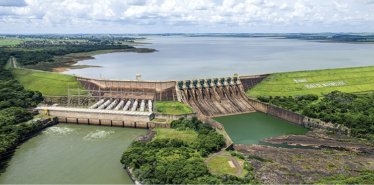 Imagem: Fotografia. Vista aérea de uma usina hidrelétrica. Na parte superior, um lago grande e extenso, em seguida, uma barragem e abaixo, dois lagos pequenos. Em volta há muitas árvores. Fim da imagem.