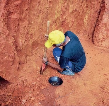 Imagem: Fotografia. Um homem está agachado e segurando uma pá sobre terra em um barranco.  Fim da imagem.