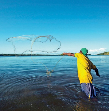 Imagem: Fotografia. Um homem está dentro de um rio e jogando uma rede de pesca na água. Fim da imagem.