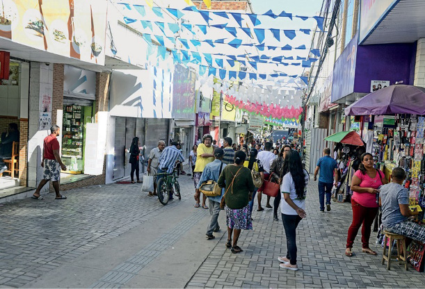 Imagem: Fotografia. Pessoas andando em uma rua com bandeirinhas coloridas penduradas. Nas laterais há lojas.  Fim da imagem.