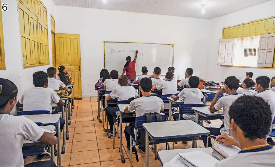 Imagem: Fotografia 6. Alunos com camiseta branca e azul estão sentados e carteiras escolares e de costas. Ao fundo, uma professora está escrevendo na lousa. Fim da imagem.