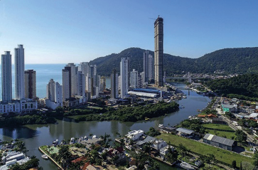 Imagem: Fotografia. Vista aérea de um rio com construções nas margens. Ao fundo, morros e o mar. Fim da imagem.