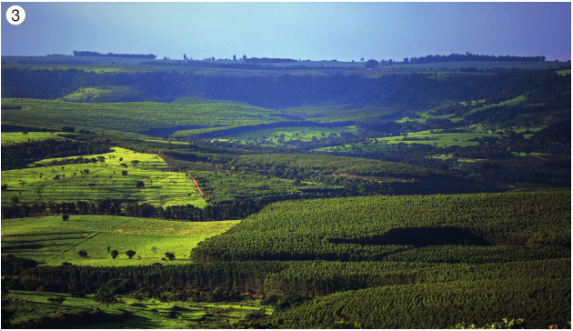 Imagem: Fotografia 3. Vista aérea de um terreno mais baixo entre dois morros verdes.  Fim da imagem.