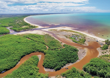 Imagem: Fotografia. Vista aérea de um rio sinuoso com água marrom. Em volta há muitas árvores e à direita, o rio se encontra com outro rio e ambos deságuam no mar. Fim da imagem.